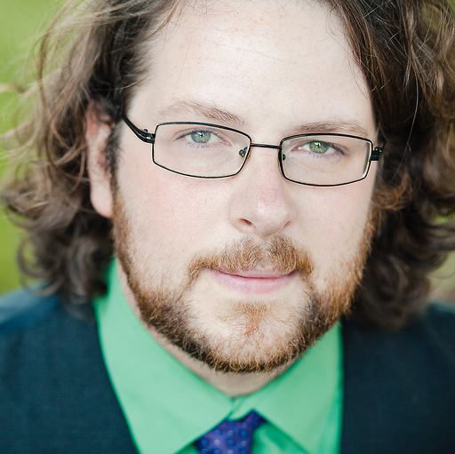 Chris, a white man with a beard and shoulder-length curly hair, looks into the camera. He is wearing glasses, a green shirt with a purple tie, and a dark blazer. 