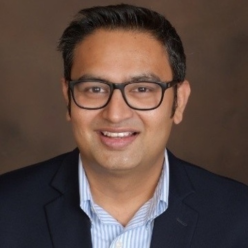 A person wearing glasses and a blazer, smiling in front of a brown background.